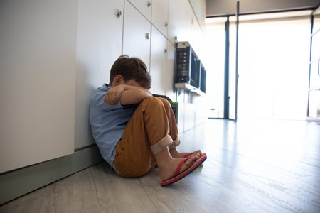 Child boy hugging knees and resting head against white cabinets in home hallway with intercom. Innocence, solitude, comfort, contemporary, minimalism, relaxation, natural lightの写真素材