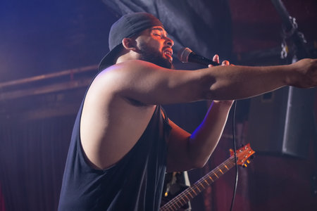 Male vocalist holding mic and extending arm under amber purple spotlights near guitar headstock. Live music, performance, concert, stage, entertainment, musician, sound equipmentの写真素材