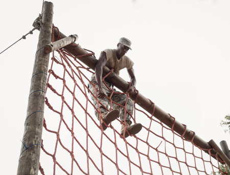 African American male soldier climbing red rope net obstacle course gripping beam and hoisting legs. Military, training, outdoor, endurance, strength, rugged, disciplineの写真素材