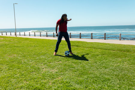 African American man preparing to kick soccer ball on grass beside coastal railing under clear sky. Athletic, leisure, outdoor, vibrant, seaside, activity, fitnessの写真素材