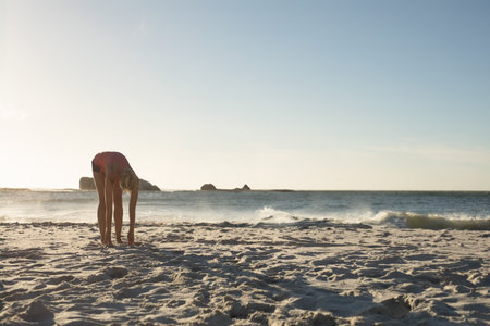 Female exerciser bending forward on beach by ocean waves in red bikini near islets, copy space. Fitness, wellness, relaxation, nature, recreation, serenity, minimalistの写真素材