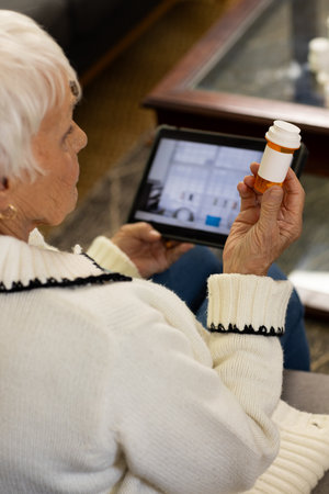 Senior woman holding prescription bottle and comparing label with tablet on sofa in living room. Elderly, wellness, contemplation, modern, domestic, health, lifestyleの写真素材