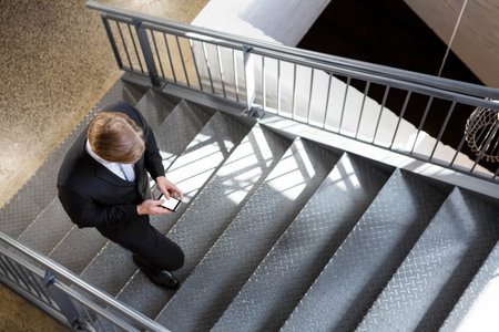 Suited businessman is descending staircase in office using smartphone, under geometric shadows. Professional, corporate, modern, urban, sleek, technology, workspaceの写真素材