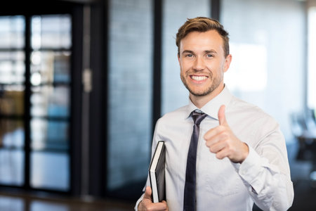 Business professional standing in office with glass panels holding two notebooks giving thumbs up. Modern, corporate, success, teamwork, innovation, professional, achievementの写真素材