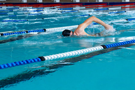 Male swimmer performing front crawl at aquatic center pool with swim cap, goggles and lane ropes. Water sport, athleticism, fitness, competition, aquatic, freestyle, movementの写真素材