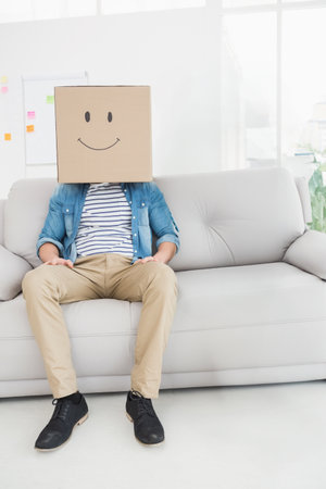 Sitting on light grey sofa in lounge, man wearing cardboard box by flipchart, copy space. Creative workspace, casual attire, playful expression, interior design, modern office, leisure environment, artistic conceptの写真素材