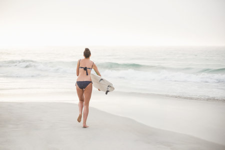 Female surfer walking toward shoreline on sandy beach in polka-dot bikini carrying white surfboard. Coastline, tranquility, leisure, adventure, minimalist, natural, outdoorの写真素材