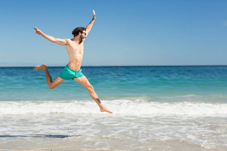 Man wearing turquoise swim shorts and dark sunglasses leaping above surf on sandy beach, copy space. Vibrant, energetic, lifestyle, outdoor, athletic, youthful, adventureの写真素材