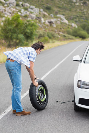 Middle-aged man lifting spare wheel beside sedan on mountain road using car jack, copy space. Automobile, adventure, outdoor, rugged, travel, repair, landscapeの写真素材