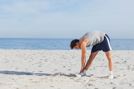 Man stretching forward in sleeveless top, sports shorts, athletic shoes on sandy beach, copy space. Fitness, wellness, outdoor, exercise, serenity, athleticism, landscapeの写真素材