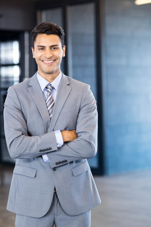 Smiling Hispanic man wearing grey suit and crossing arms in office corridor with glass partitions. Professional, corporate, modern, sleek, confident, business, executiveの写真素材