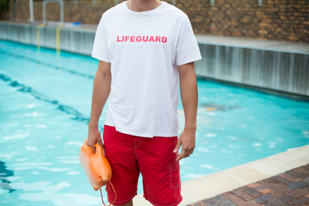 Lifeguard standing at pool edge holding orange rescue buoy beside turquoise water and brick deck. Rescue, safety, aquatic, vigilance, sport, urban, outdoorの写真素材