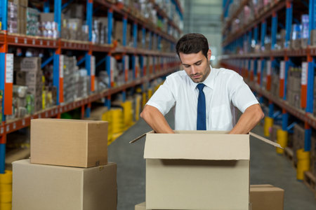 Cardboard boxes sitting on table amid tall metal shelving units in large warehouse aisle. Storage, logistics, industrial, shipping, inventory, organizational, supply chainの写真素材