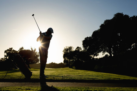 Mature adult male golfer swinging club on tee at golf course with golf bag, copy space. Athletic, leisure, landscape, outdoor, sport, elegance, adventureの写真素材