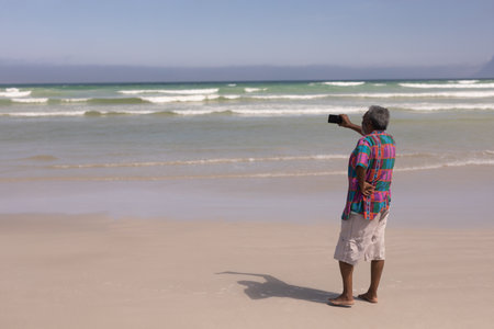 Senior man standing barefoot on sandy shoreline capturing ocean view with smartphone, copy space. Landscape, leisure, nature, tranquility, vibrant, outdoor, explorationの写真素材