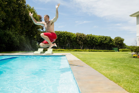 Adult male jumping into clear blue pool at backyard deck with white lounge chairs, copy space. Athleticism, leisure, outdoor, relaxation, serenity, lifestyle, recreationの写真素材