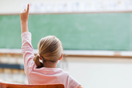 Female child student sitting at wooden desk raising hand in classroom before large green chalkboard. Education, learning, classroom, school, student, teaching, academicの写真素材