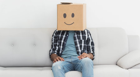Adult male sitting on light grey sofa against white wall, wearing plaid shirt, cardboard box mask. Minimalist, eccentric, playful, quirky, contemporary, expressive, casualの写真素材