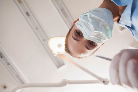 Female dentist wearing mask holding probe in clinic under overhead lamp and ceiling panels. Clinical, healthcare, sterile, professional, examination, hygiene, medicalの写真素材