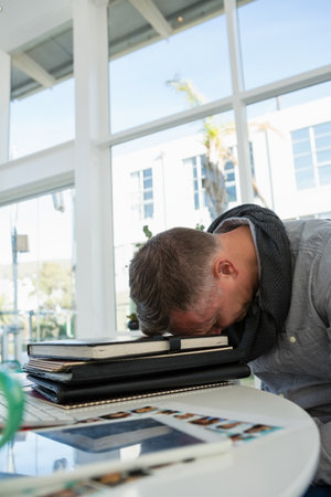 Middle-aged man resting head on notebooks at round white table in coworking space, copy space. Concentration, productivity, workspace, modern, reflective, creative, urbanの写真素材
