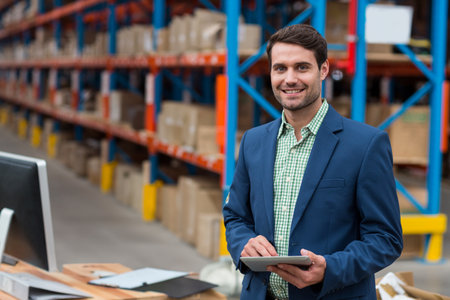 Tablet sitting on table beside paperwork and monitor in warehouse aisle lined with cardboard boxes. Industrial, logistics, supply chain, inventory, organization, operational, modernの写真素材