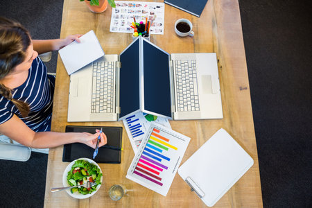 Indian woman working at office workspace while sketching on tablet with laptops, coffee, copy space. Creative workspace, professional environment, digital artistry, productivity tools, modern office, artistic process, office decorの写真素材