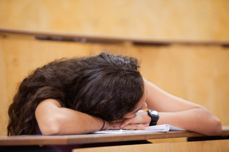 female student resting head on arms atop open notebook with digital wristwatch on desk in classroom. Learning, education, study, concentration, academic, introspection, minimalismの写真素材