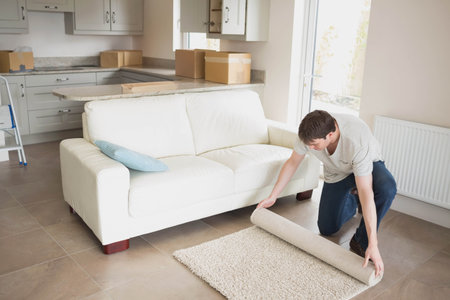 Man kneeling on tiled floor unrolling small rug beside white sofa in living room, copy space. Interior, home staging, renovation, minimalism, casual, bright, functionalの写真素材