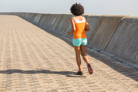 African American woman jogging along sea wall path wearing orange top and running shoes, copy space. Athletic, outdoors, fitness, movement, leisure, urban, vibrantの写真素材