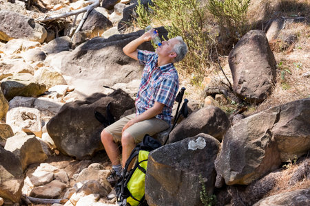 Mature man sitting on boulder drinking water from bottle with backpack, hiking poles on creek bed. Adventure, exploration, outdoor, wilderness, tranquility, nature, ruggedの写真素材