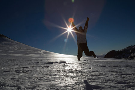 Male jumping into air wearing padded winter jacket, ski goggles on snowy mountain under sun flare. Adventure, outdoor, exploration, extreme, active, rugged, scenicの写真素材
