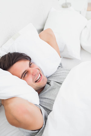 Man wearing gray t-shirt lying on bed and smiling in bedroom with pillows and lamp base. Relaxation, comfort, sleep, cozy, tranquility, home interior, wellnessの写真素材