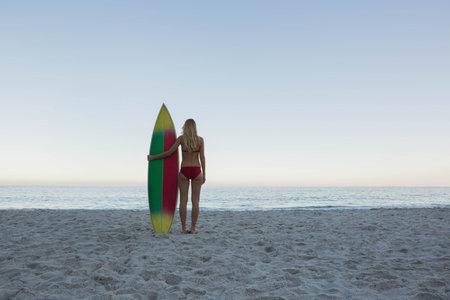 Woman standing barefoot on sandy beach at dawn facing ocean horizon and holding striped surfboard....