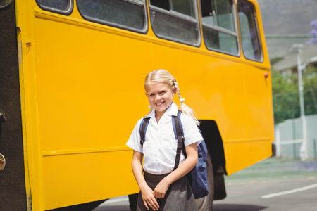 Female child student standing at school bus parking area beside yellow bus with dark blue backpack. Vibrant, youthful, outdoor, educational, cheerful, playful, adolescentの写真素材
