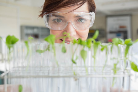 Female scientist peering over metal test tube rack with seedlings in lab wearing coat and goggles. Botany, innovation, experimentation, growth, science, biotechnology, researchの写真素材