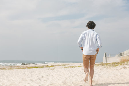 Man walking barefoot toward ocean waves on beach by boardwalk ramp, copy space. Seascape, relaxation, leisure, tranquility, nature, wellness, outdoorの写真素材