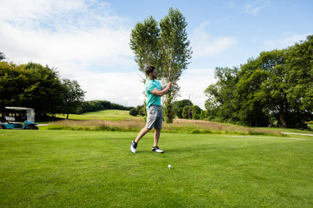Man swinging golf club on tee box at golf course overlooking fairway with ball and cart. Sport, leisure, outdoor, health, activity, nature, recreationの写真素材