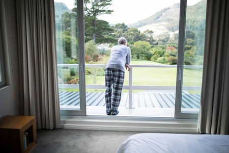 Senior man wearing pajamas leaning on balcony railing gazing at manicured lawn through glass doors. Contemplation, tranquility, landscape, leisure, serenity, comfort, outdoorの写真素材
