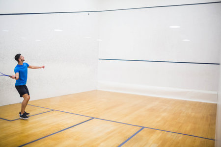 Male player is standing on wooden squash court under overhead lights holding racket ready to strike. Athletic, sport, active, competitive, fitness, movement, trainingの写真素材