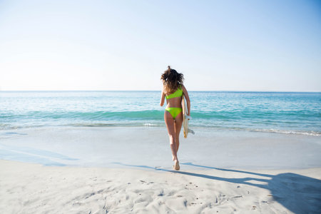 Woman walking on sandy beach carrying surfboard while wearing green swimsuit toward calm blue ocean. Coastline, outdoor, vibrant, leisure, adventure, relaxation, minimalismの写真素材
