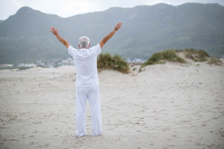 Senior man standing on wide sandy shoreline near dunes facing coastal town under overcast sky. Seascape, tranquility, solitude, contemplative, minimalist, natural, outdoorの写真素材