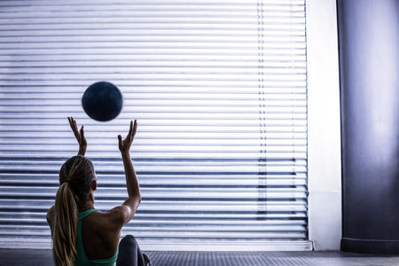 Woman wearing green tank top tossing black medicine ball on mat by shutter door in gym. Fitness, strength, workout, training, resilience, athletic, determinationの写真素材