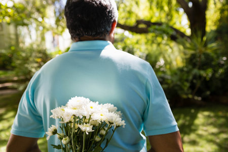 Senior man wearing blue polo shirt holding white daisy bouquet behind back in backyard. Romance, tranquility, nature, contemplation, leisure, outdoor, freshnessの写真素材