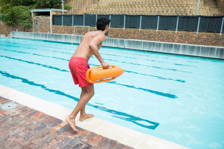 Mid-adult male lifeguard standing poolside holding rescue buoy by lanes in red trunks, copy space....