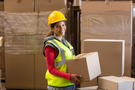 Female warehouse worker holding medium cardboard box in storage warehouse, with stacked pallets....