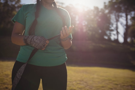 Female athlete adjusting patterned hand wraps in sunlit grassy park area with lens flare. Fitness,...