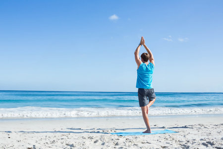 Athletic man in sportswear balancing on yoga mat on sandy beach with fitness tracker, copy space....