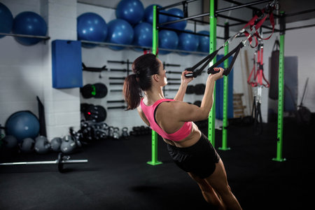 Female fitness trainee gripping black suspension straps and pulling body at gym functional rig area....