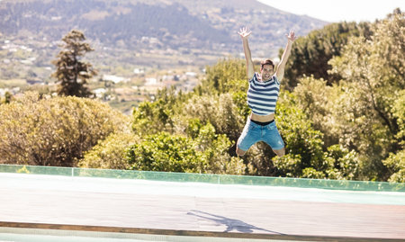 Man jumping above wooden pool deck with glass barrier, overlooking green valley and terraced hills. Adventure, leisure, outdoor, freedom, dynamic, scenic, vitalityの写真素材