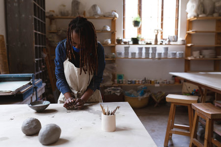 African American woman kneading clay at ceramics workshop table wearing apron with pottery tools. Artisan, crafting, creativity, studio, handmade, artisanal, rusticの写真素材